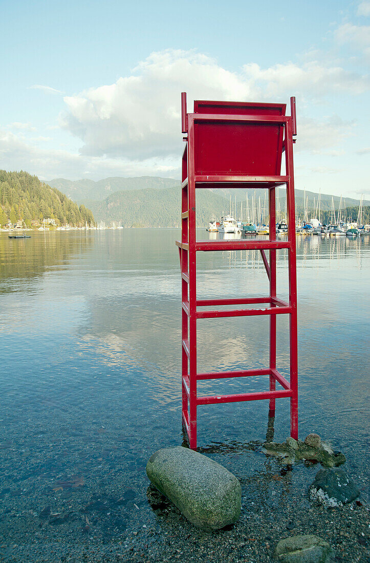 A tall empty wooden lifeguard chair painted red,  overlooking the clear calm waters of Deep Cove. , Indian Arm in Deep Cove, North Vancouver, BC, Canada