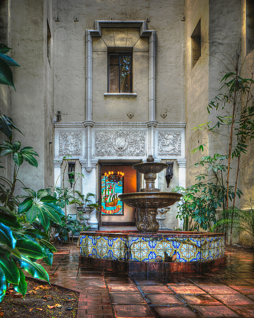 The courtyard of the Villa Rosa apartments building, with a small stone fountain and pool.  Spanish style glazed tiles around the centrepiece. Door and a stained glass window. Plants. Night. Dusk. Retro style architecture and decor., Los Angeles, Californ