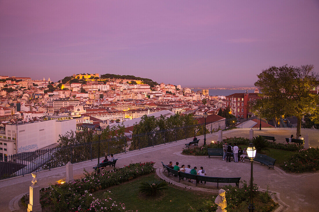 Menschen auf Parkbänken am Aussichtspunkt Miradouro Sao Pedro de Alcantara im Stadtviertel Chiado mit Blick auf die Baixa Unterstadt und das Castelo de San Jorge in der Abenddämmerung, Lissabon, Portugal