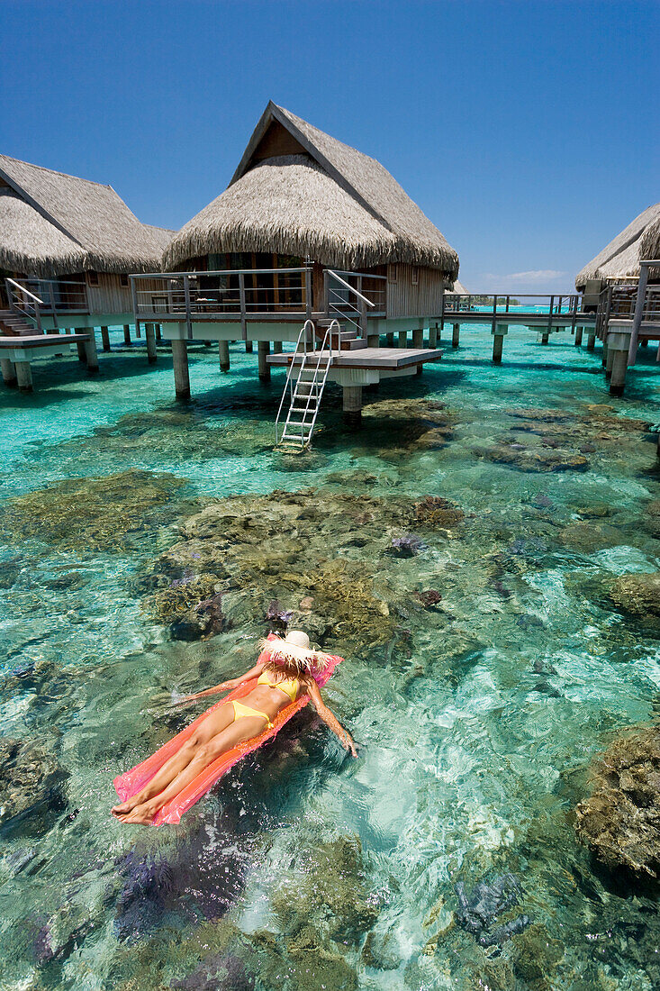 French Polynesia, Moorea, Woman sunbathing on inflatable raft over ocean reef, Luxury resort bungalows over ocean.