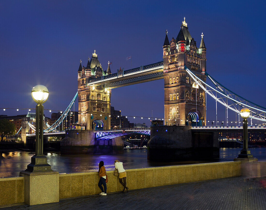 Two women looking at Thames, Tower Bridge with lights at night, London, England