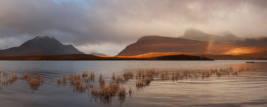 Blick über den Lochan an Ais auf die Gipfel des Inverpolly Nature Reserve mit einem Regenbogen über dem See, Ullapool, Sutherland, Schottland, Großbritannien