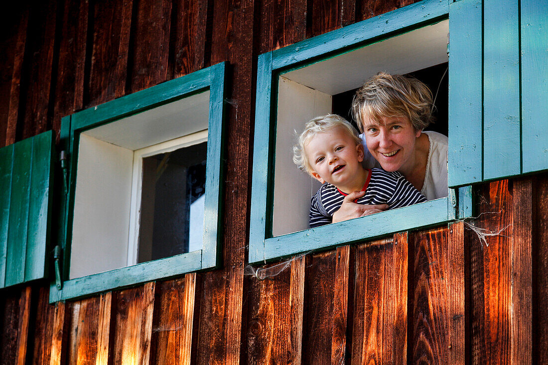 Mutter und Sohn (2 Jahre) schauen aus einer Holzhütte, bei Maria Alm, Pinzgau, Salzburger Land, Österreich