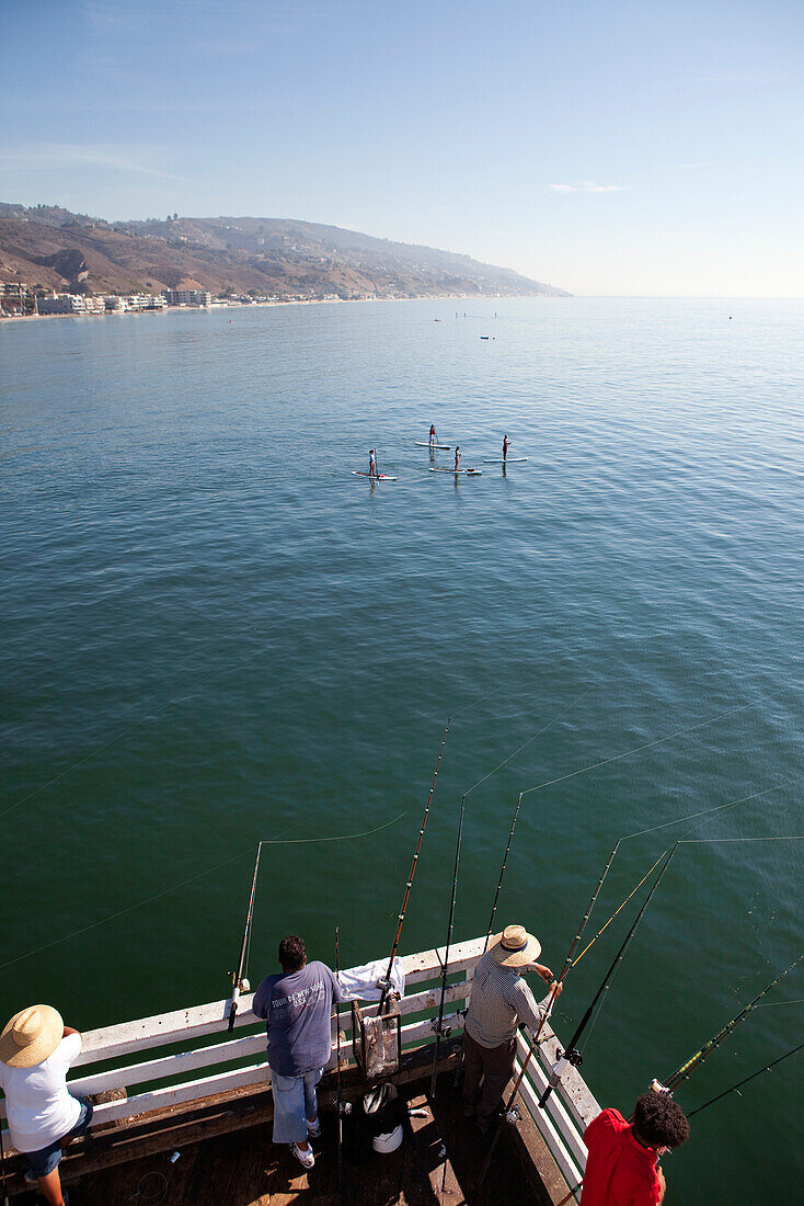 USA, California, Malibu, men fish off of the Malibu pier while four paddleboarders pass by on calm seas