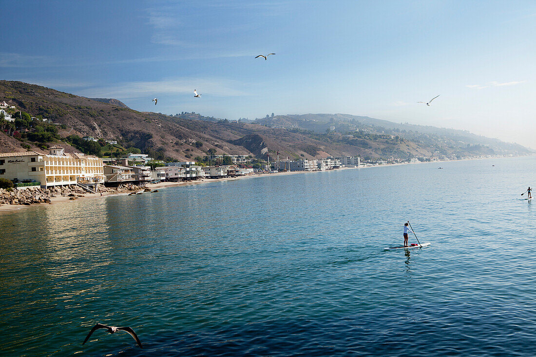 USA, California, Malibu, paddleboarders cruise by the Malibu pier with the Malibu Beach Inn in the distance