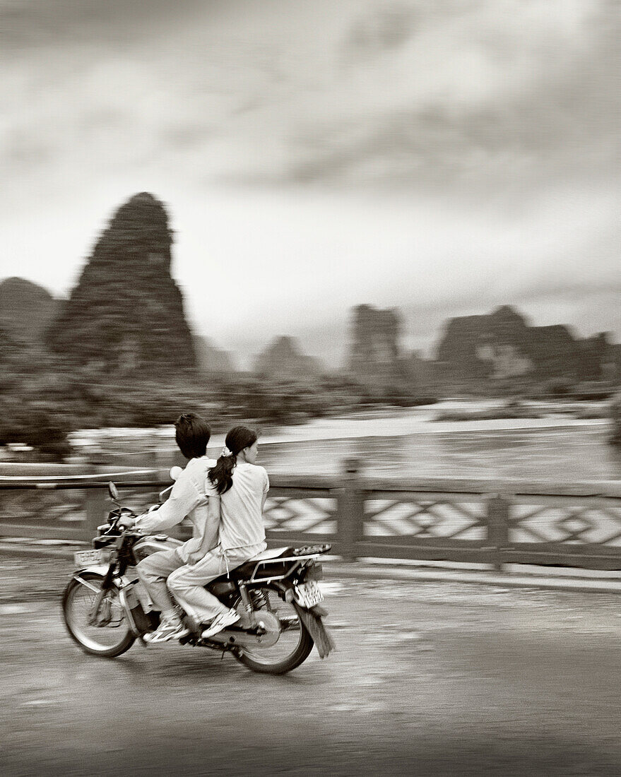 China, Guilin, people traveling in motorbikes on bridge with mountains in the background (B&W)