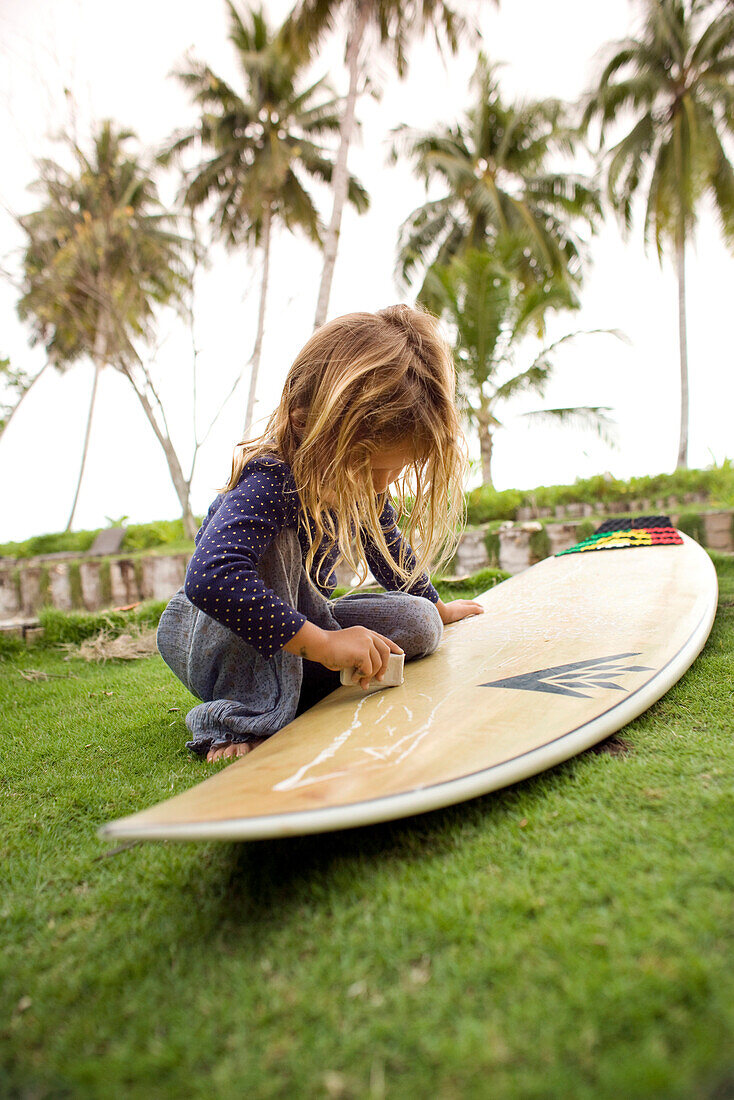 INDONESIA, Mentawai Islands, Kandui Surf Resort, girl waxing surfboard on lawn with palm trees in the background