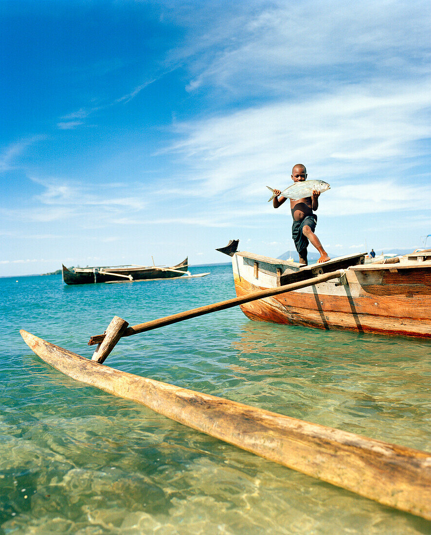 MADAGASCAR, boy holding fish standing on dugout canoe, Nosy Komba