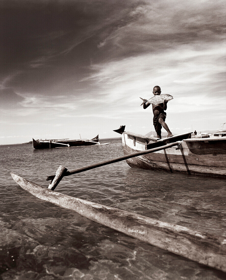 MADAGASCAR, Nosy Komba, Jardin Vanille, boy holding a Travali fish standing on an outrigger canoe, Indian Ocean