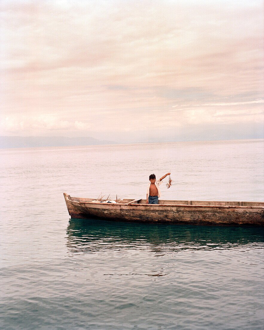 PANAMA, Bocas del Toro, a boy sitting in a large dugout canoe sits holding up a lobster and waits for his dad to surface with more food, the Caribbean Sea off the coast of Isla Colon, Central America