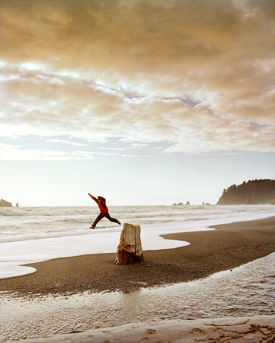 USA, Washington State, man jumping off of a tree stump at Rialto Beach, Olympic National Park