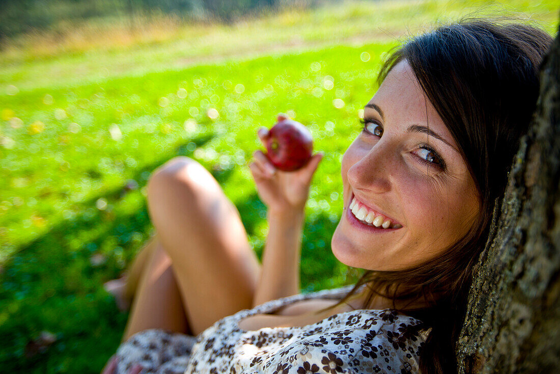 Junge Frau mit einem Apfel lehnt an einem Baumstamm, Steiermark, Österreich