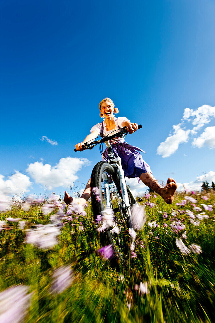 Junge Frau im Dirndl fährt mit einem Mountainbike über eine Wiese, Duisitzkar, Planai, Steiermark, Österreich
