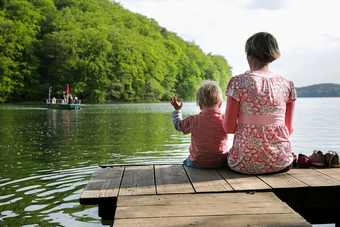 Mutter und Sohn sitzen auf einem Steg am Schmalen Luzin, Luzinfähre im Hintergrund, Naturpark Feldberger Seenlandschaft, Mecklenburg-Vorpommern, Deutschland