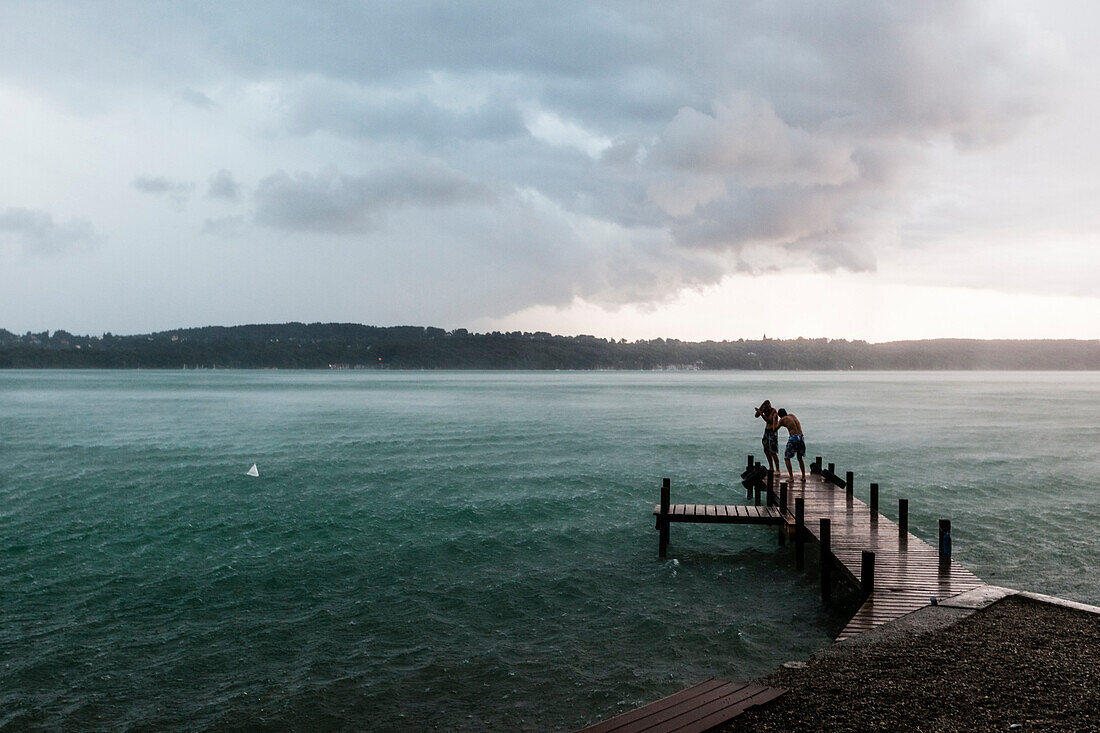 Two young men standing on a jetty at Lake Starnberg in rain, Bavaria, Germany