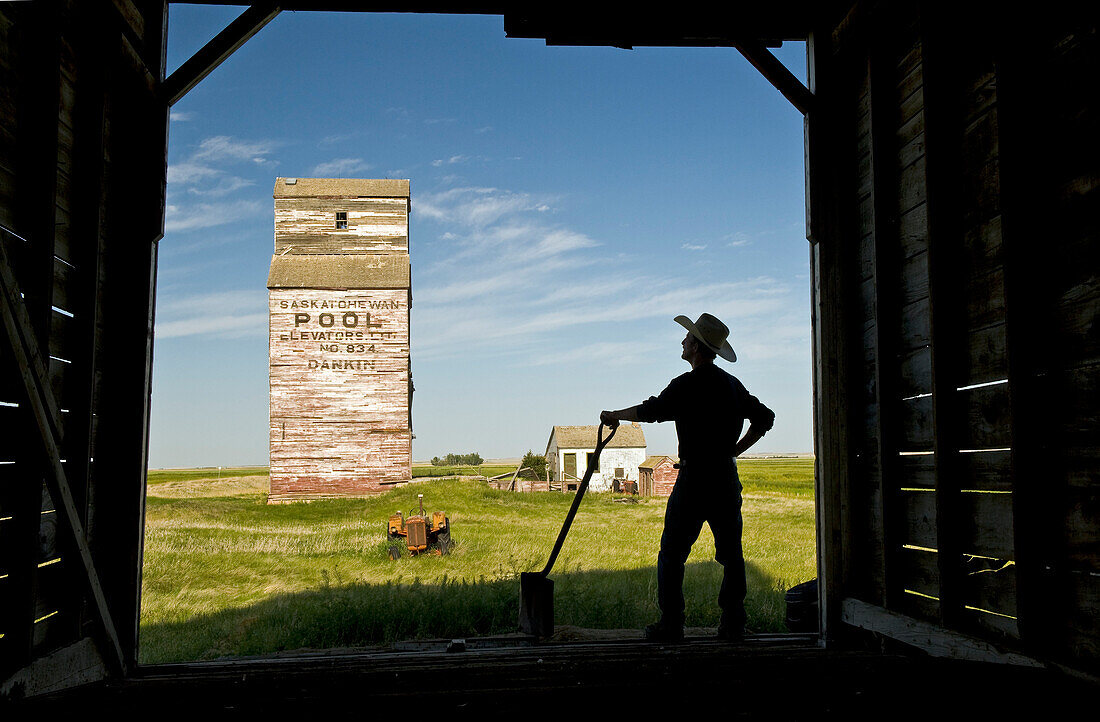 Farmer with a shovel looking out at an abandoned grain elevator, Dankin, Saskatchewan