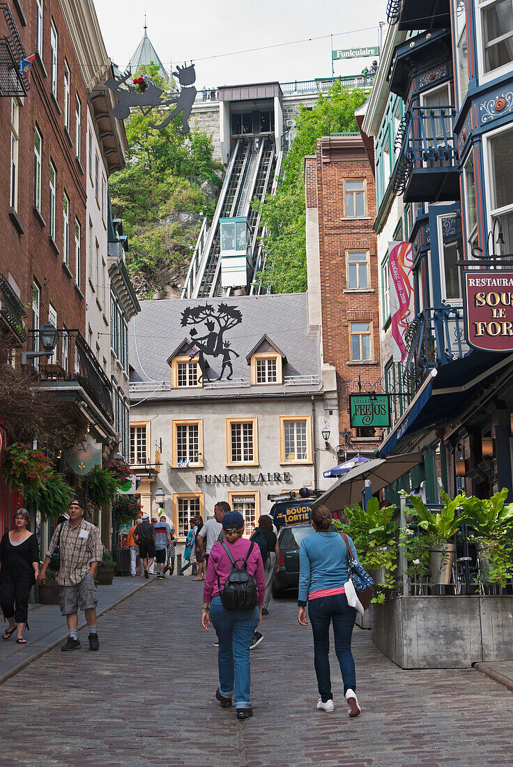 Two women walk up cobblestone streets past shops toward a funicular as car ascends, quebec city quebec canada
