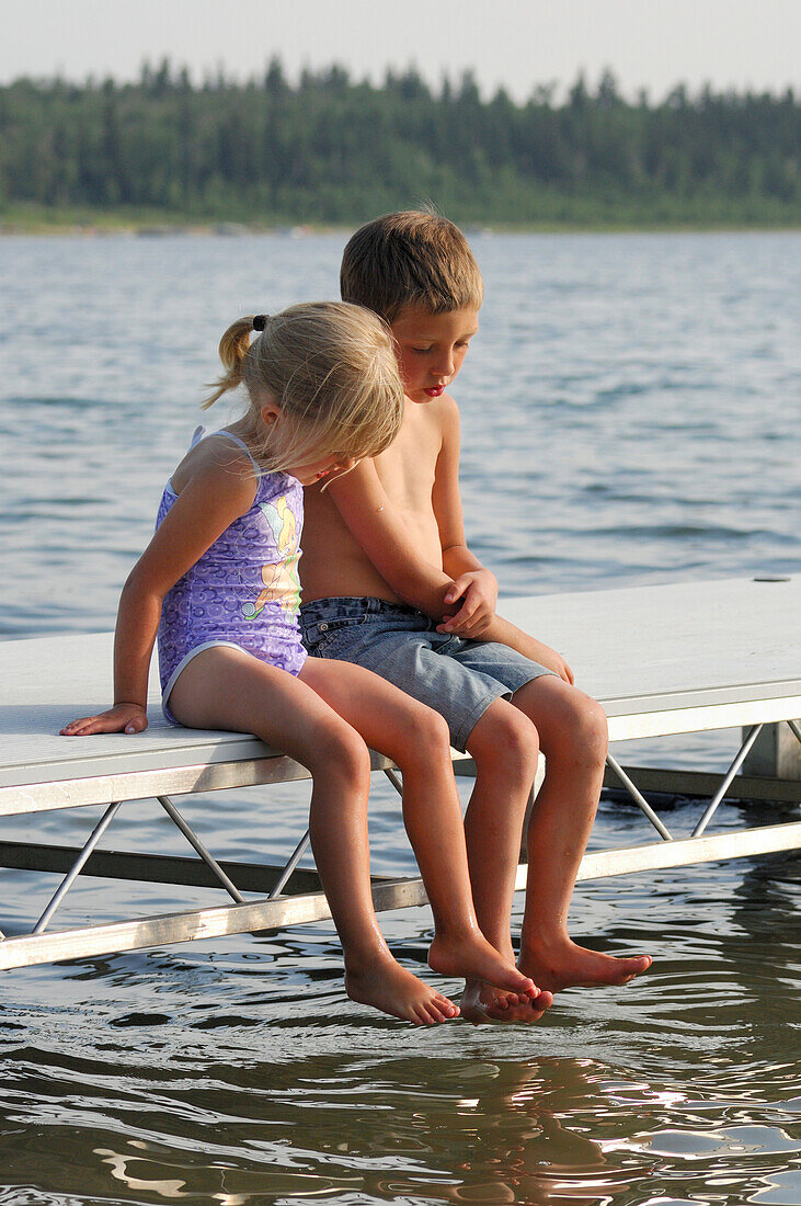 Brother and sister sitting on a lake dock looking into the water, Lac Sante, Alberta