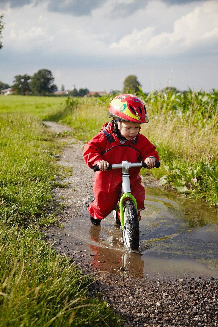 Boy with balance bicycle passing a puddle, Murnau, Bavaria, Germany