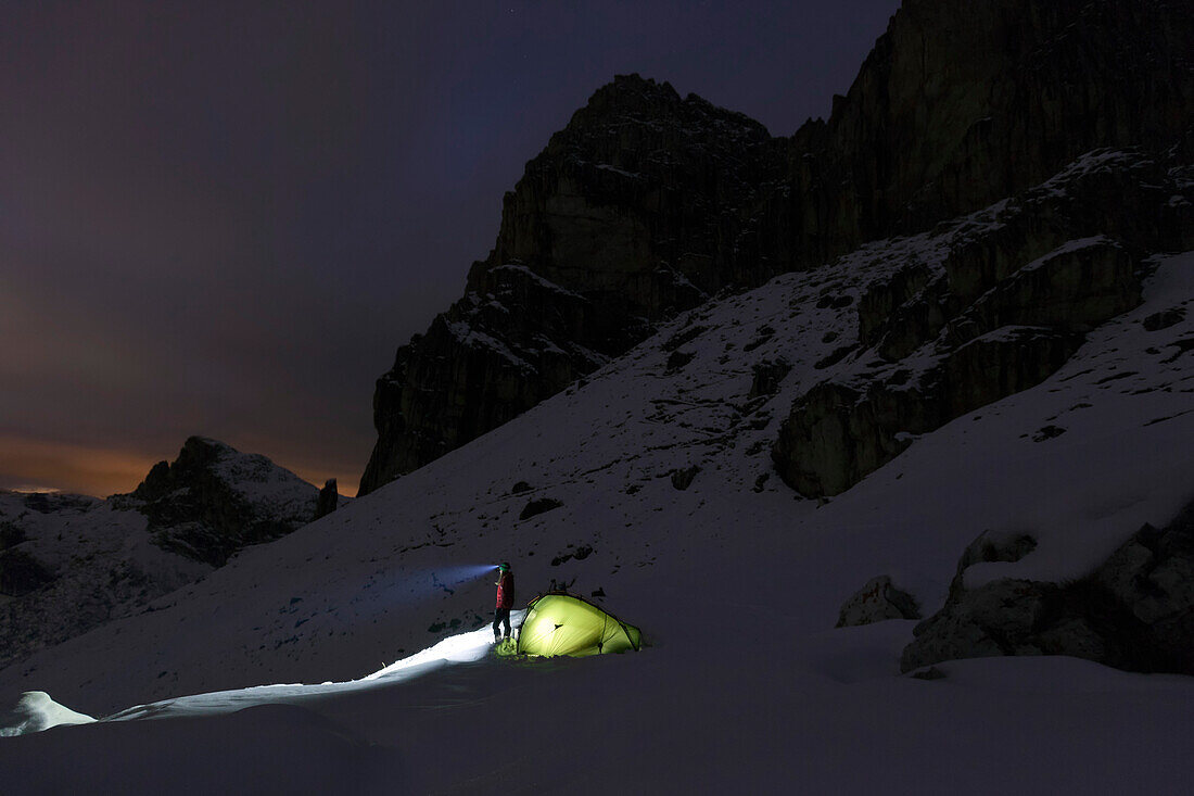 Junge Frau mit Stirnlampe steht neben einem Zelt im Schnee, Dolomiten, Belluno, Venetien, Italien