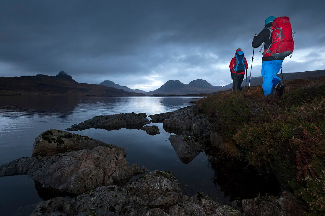 Young couple with backpacks hiking at Loch Bad a Ghaill, Stac Pollaidh, Cul Beag, Sgorr Tuath and Ben Mor Coigach in background, Assynt, Scotland, United Kingdom