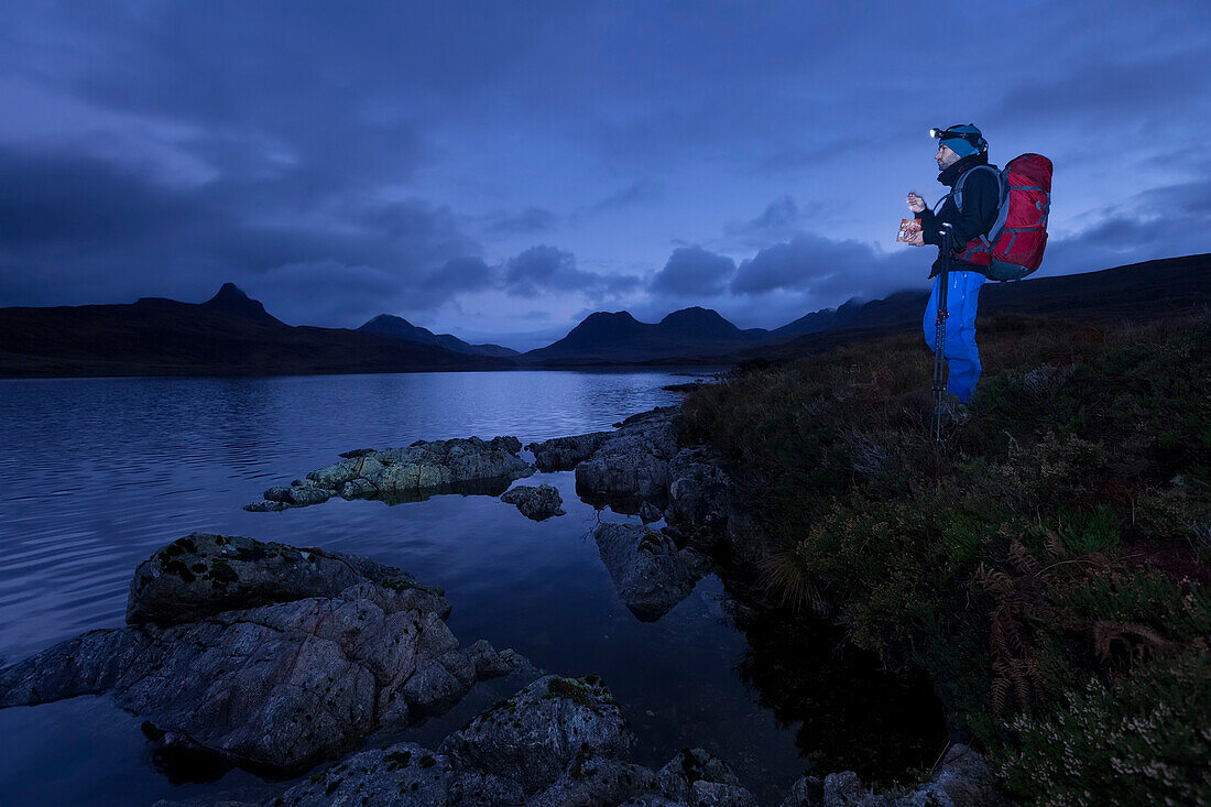 Young man standing at Loch Bad a Ghaill at dusk, Stac Pollaidh, Cul Beag, Sgorr Tuath and Ben Mor Coigach in background, Assynt, Scotland, United Kingdom