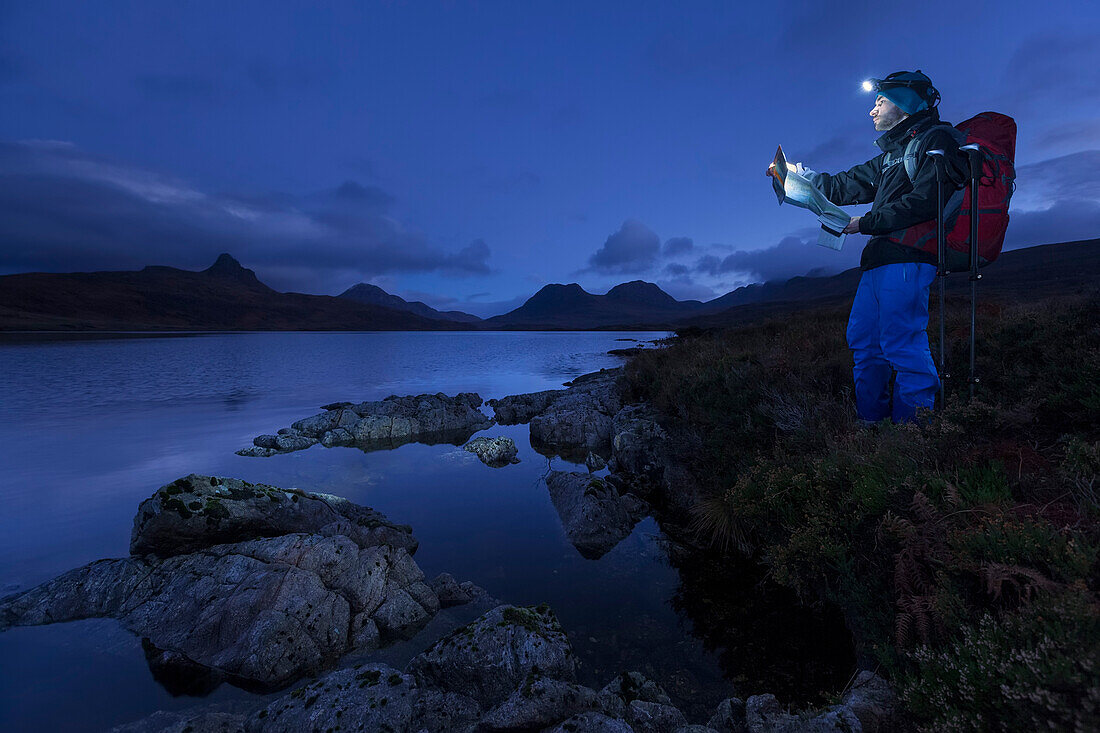 Junger Mann mit Stirnlampe liest eine Wanderkarte am Loch Bad a Ghaill in der Dämmerung, Stac Pollaidh, Cul Beag, Sgorr Tuath und Ben Mor Coigach im Hintergrund, Assynt, Schottland, Großbritannien