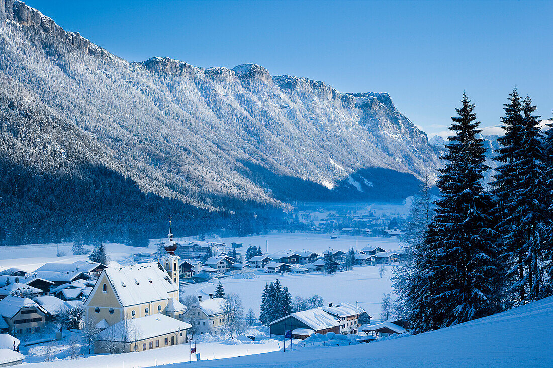 Snow Covered Village With Mountains And Picturesque Church In A Valley Below Ski Slopes At Waidring, Austrian Alps, Austria