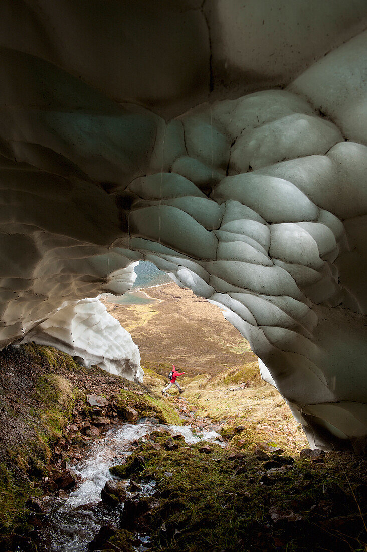 Walker Jumping Over Small Stream Coming Out Of Ice Cave, Gleann Einich Near Aviemore, Highlands, Scotland, Uk