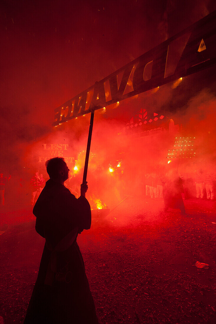 Silhoutte Of Man From Southover Bonfire Society With 'advance' Banner In Front Of Red Flares At 'bonfire Prayers' At End Of Bonfire Night, Lewes, East Sussex, Uk