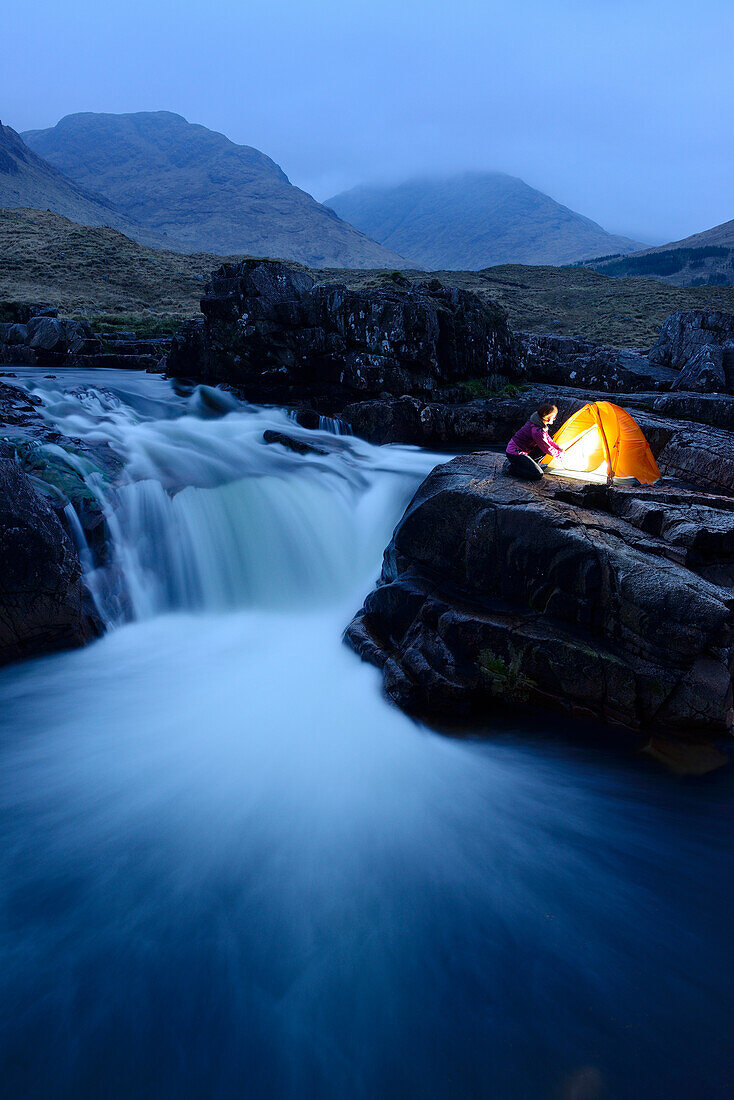 Woman sitting infront of an illuminated tent, Glen Etive, Highlands, Scotland, United Kingdom