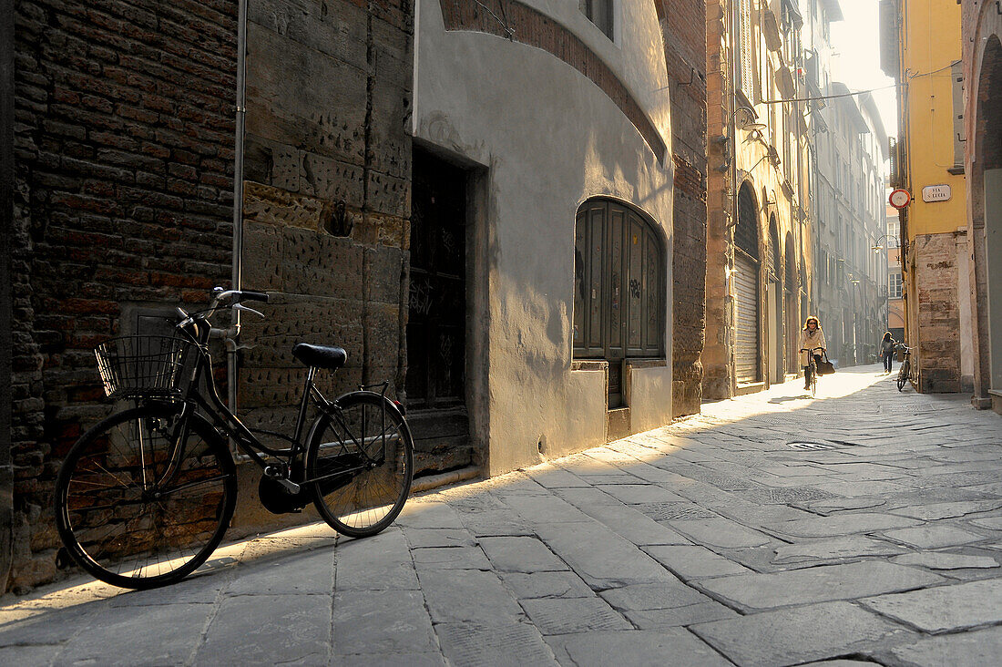 Fahrrad und Radfahrerin im morgendlichen Streiflicht in einer Gasse von Lucca, Toskana, Italien