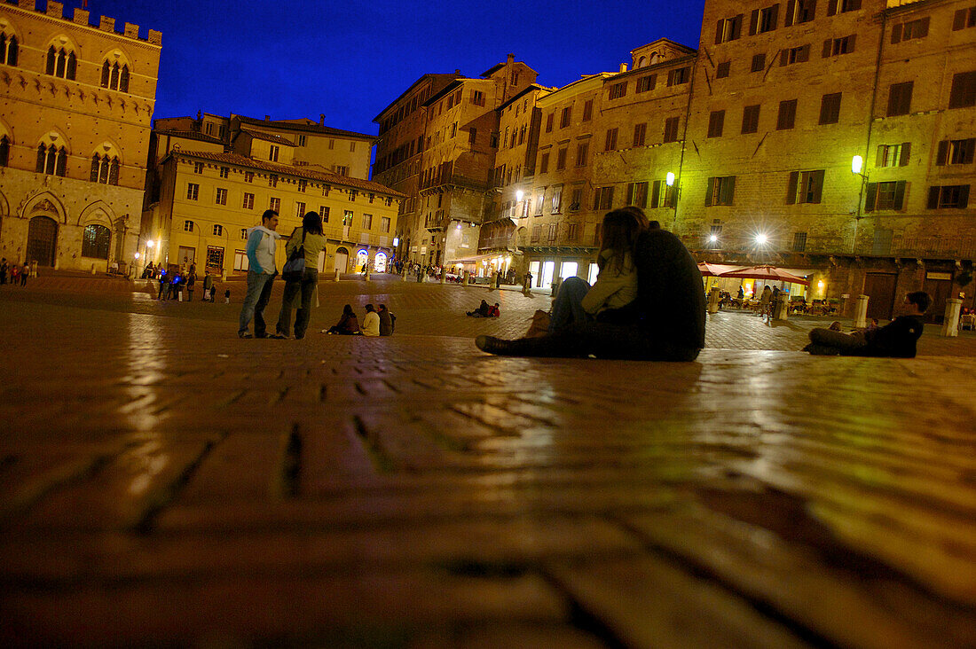 Visitors sitting on the cobbles of the Piazza del Campo at the Palazzo Pubblico, Siena, Tuscany, Italy