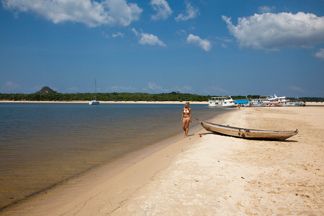 Woman strolling along the beach on a side arm of the Amazon river with Amazon river boats in the distance, Alter do Chao, Para, Brazil