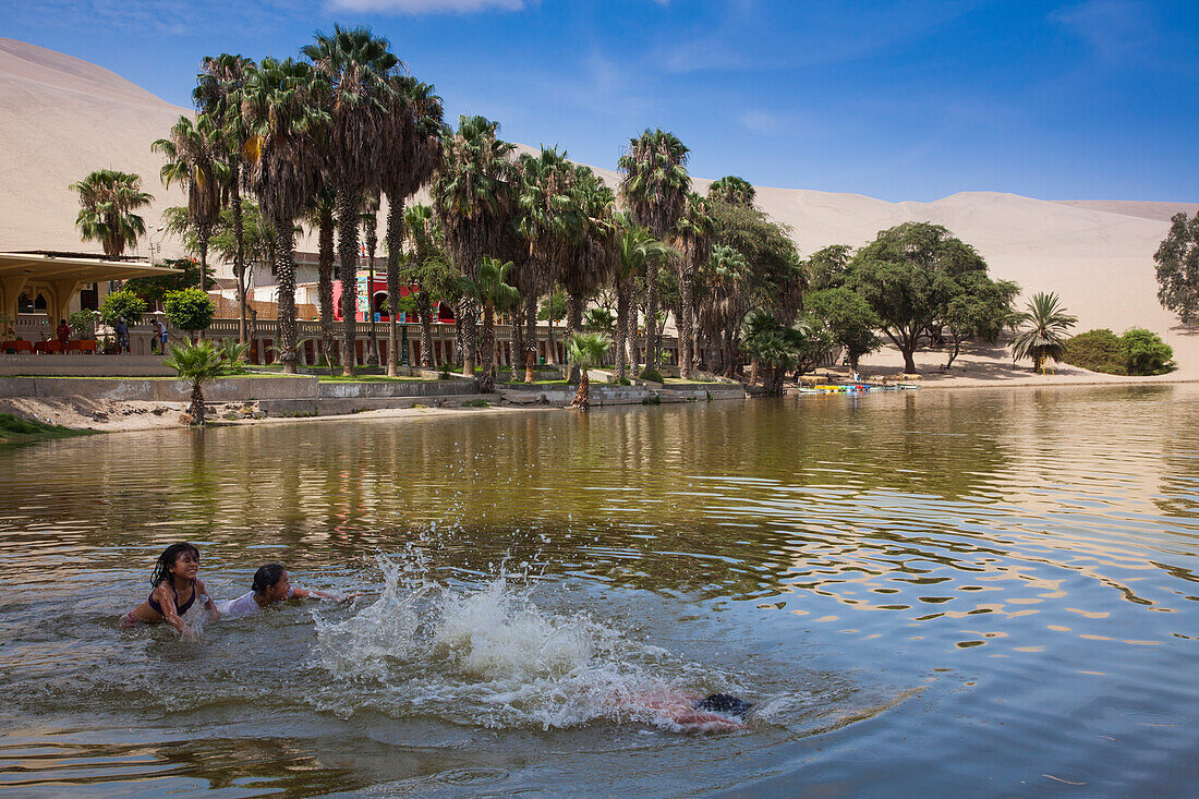 Kinder planschen im See an der Oase Huacachina mit Palmen und Sanddünen der Wüste Atacama im Hintergrund, Pisco, Ica, Chile, Südamerika