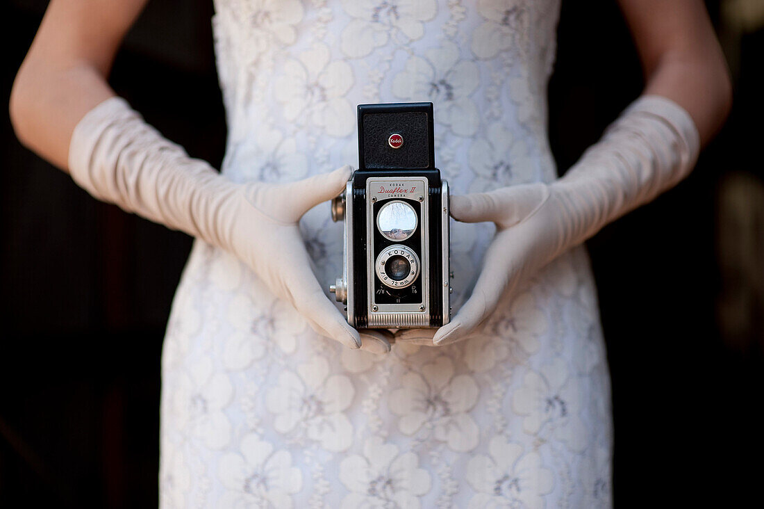 Frau in weißem Kleid und langen weißen Handschuhen sitzt im Schaukelstuhl und hält eine antike Kamera