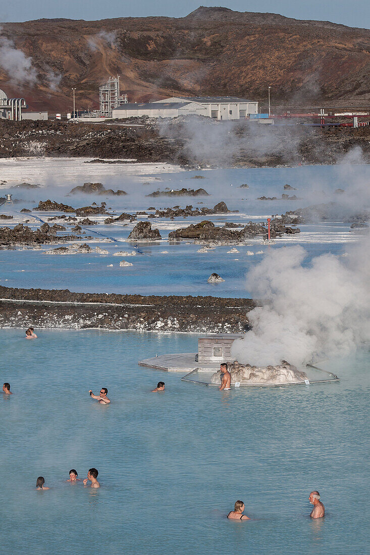 Bathers Enjoying The Hot Water Of The Blue Lagoon With, In The Background, The Svartsengi Geothermal Plant, Hot Springs And Silica Mud, Grindavik, Reykjanes Peninsula, Iceland
