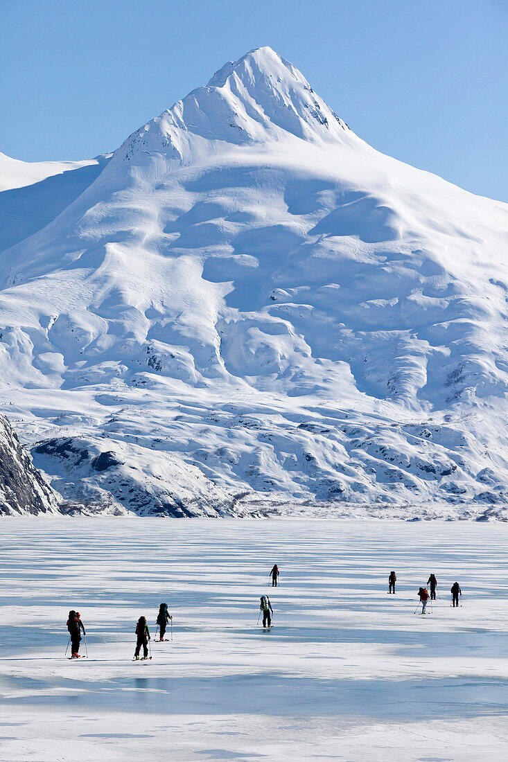 A Group Of Skiers Ski Across Frozen Portage Lake With Mount Bard In The Background, Southcentral Alaska, Spring