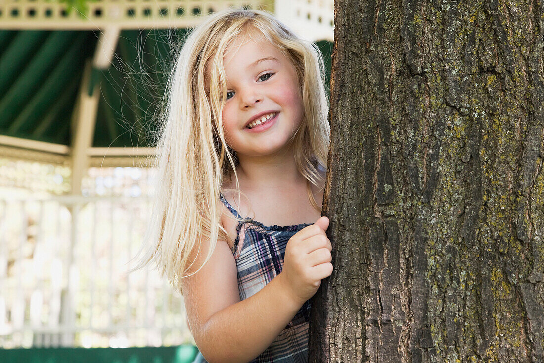 'Young Blonde Girl Smiling And Looking Out From Behind A Tree;Ontario Canada'