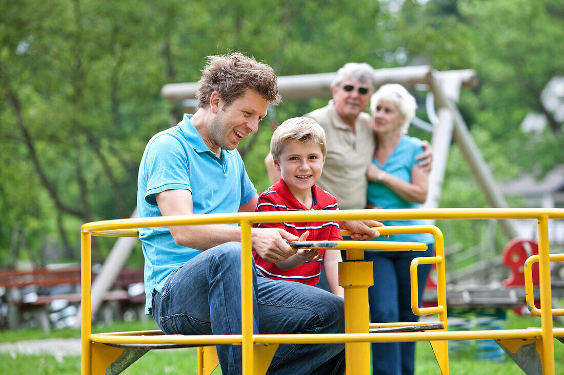 Fahter and son (7 years) at playground, grandparents in background, Styria, Austria