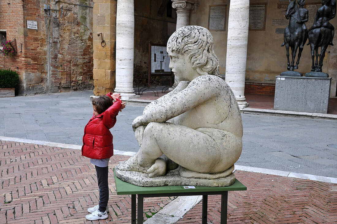 Young girl standing next to a statue on the cathedral square, Pienza, Siena, South Tuscany, Tuscany, Italy