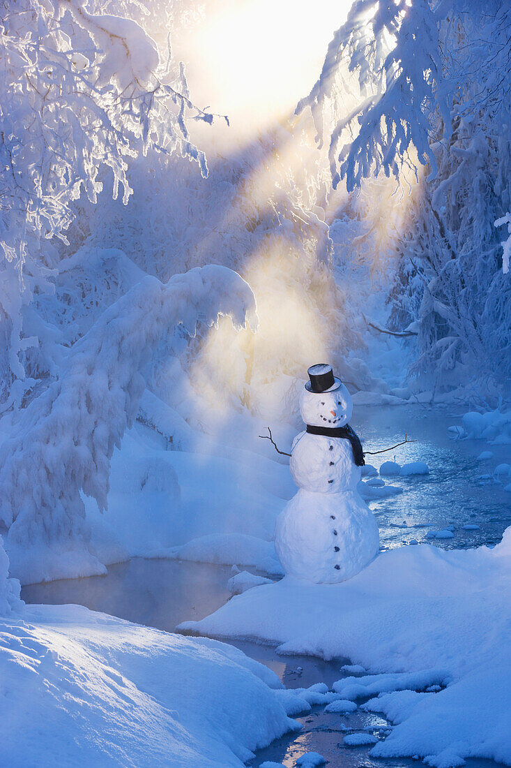 Snowman standing next to a stream with sunrays shining through fog and hoar frosted trees in the background, Russian Jack Springs Park, Anchorage, Southcentral Alaska, Winter. Digitally enhanced.