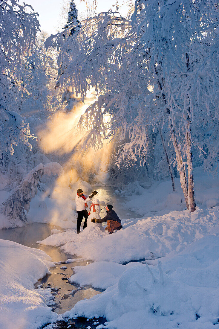 Husband and wife building a snowman in a frosty forest and backlit by sunrays, Russian Jack Springs Park, Southcentral Alaska, Winter