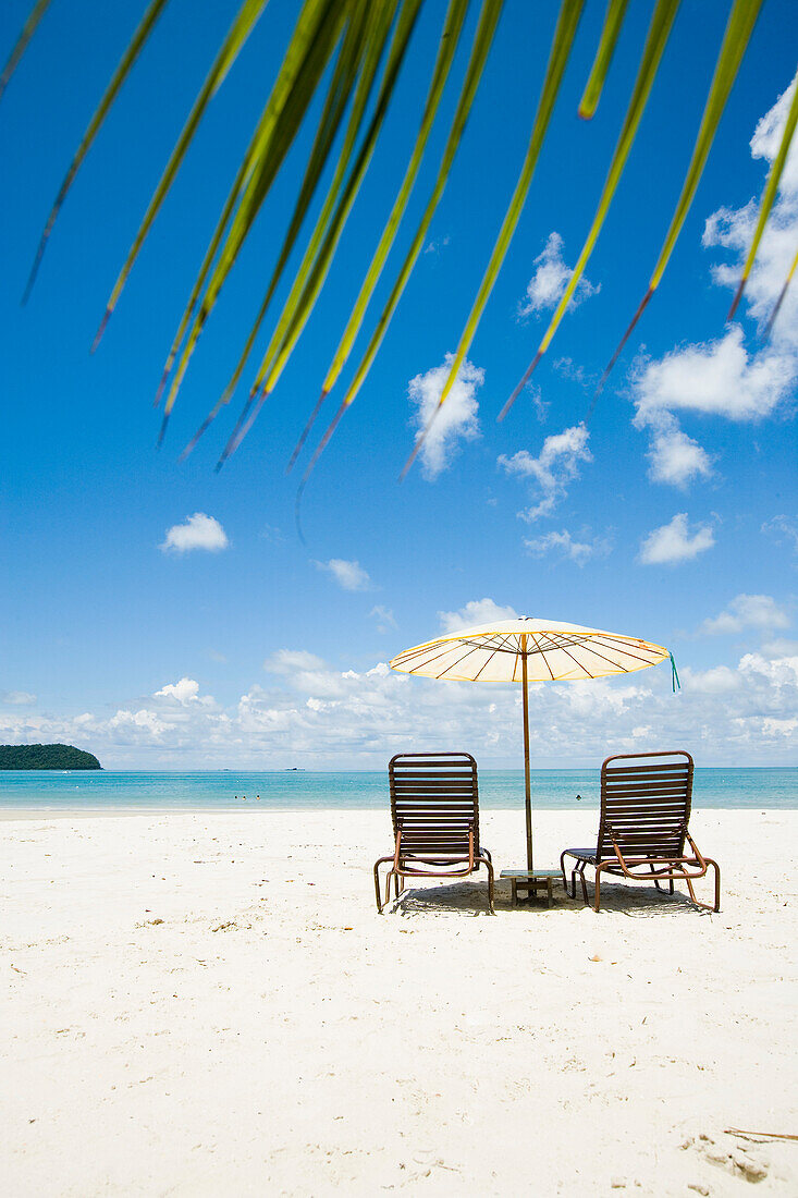 Deck chairs under parasol on white sandy beach with palm trees overlooking blue sea, Pantai Cenang (Cenang beach), Pulau Langkawi, Malaysia