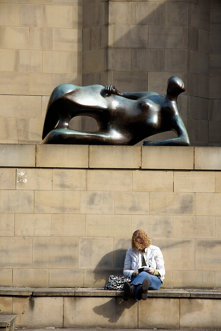 United Kingdom, England, West Yorkshire, Henry Moore sculpture outside Leeds Art Gallery and woman reading book beneath, Leeds