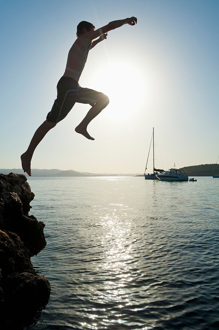 Man jumping off rocks into sea beside Cala Bassa Beach, Ibiza, Spain