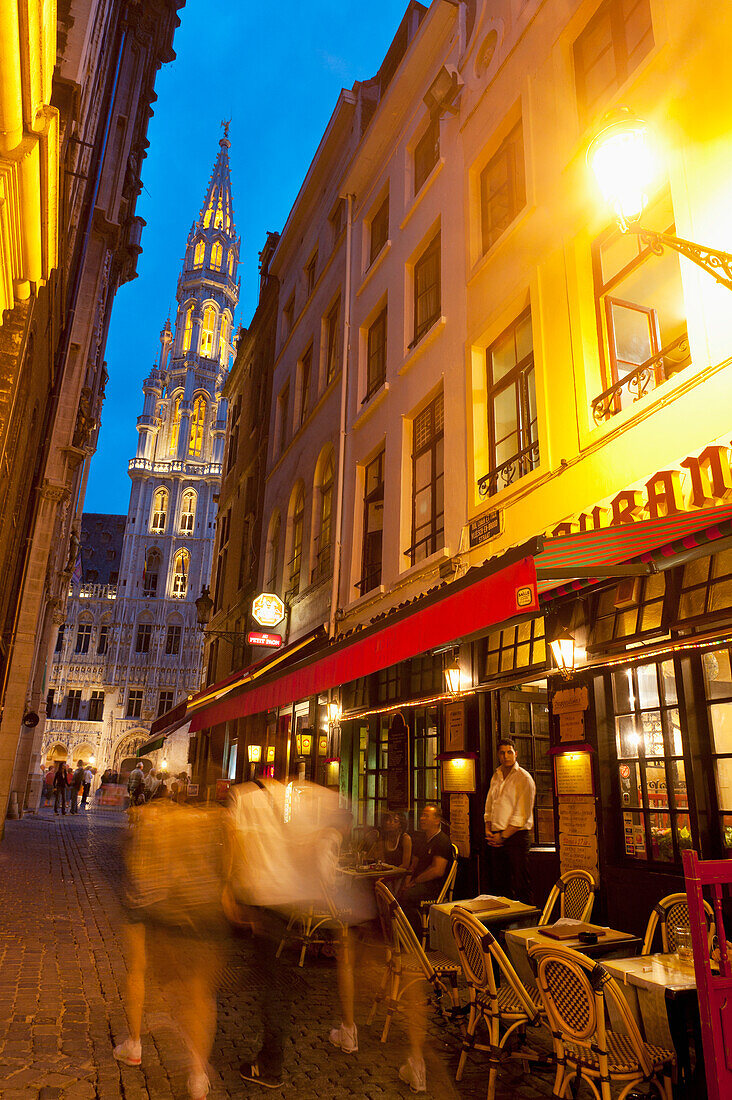 People walking down side street in front of restaurant with Town Hall behind in Grand Place at dusk, Brussels, Belgium