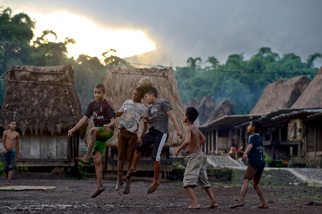 Kinder springen zum Kopfball beim Fußball, in einem Dorf der Ngada bei Bajawa, im Hintergrund traditionelle Häuser am Dorfplatz, Flores, Nusa Tenggara Timur, Östliche Kleine Sundainseln, Indonesien, Asien