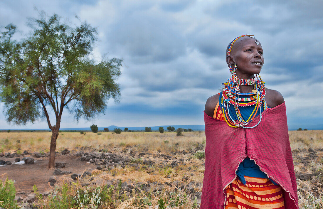 Kenya Africa Amboseli Maasai tribe village Masai woman in red costume dress and beads and tree in remote area of Amboseli National Park safari 1