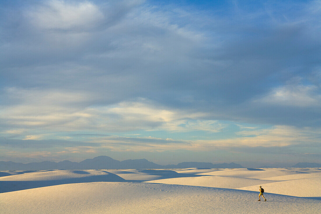 Man hiker wearing shorts, a hat and a backpack, 52, walking alone in a big sprawling landscape of sand and distant mountains, across white sand dunes, evening, autumn, White Sands National Monument, New Mexico